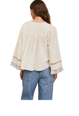 A woman shown from the back wearing a cream-colored, long-sleeve blouse with lace trim on the cuffs and neckline, paired with light blue jeans, standing against a rustic textured wall.