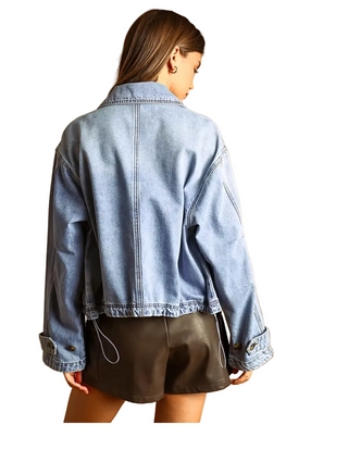 A woman shown from the back wearing a light-wash blue cropped denim jacket with rolled cuffs and a drawstring detail, paired with dark shorts, against a warm tan studio background.