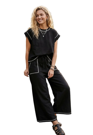 A woman models a black two-piece lounge set with white contrast stitching, featuring a short-sleeve top and wide-leg pants, set in an outdoor patio environment.