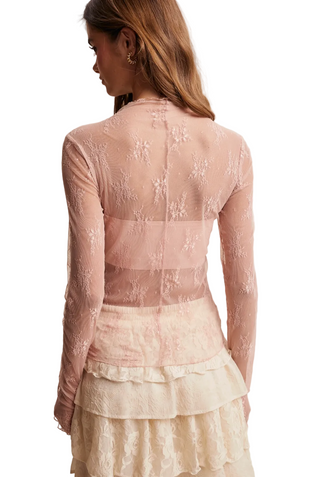 A woman shown from the back wearing a sheer blush pink long-sleeve lace top and a cream ruffled skirt against a plain beige studio background.