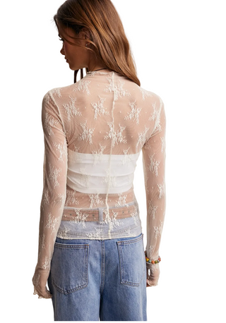A woman wearing a sheer ivory  long-sleeve top with floral lace embroidery over a bandeau-style underlayer, paired with a a denim jeans