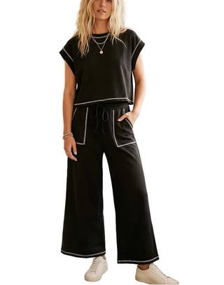 A woman models a black two-piece lounge set with white contrast stitching, featuring a short-sleeve top and wide-leg pants, set in an outdoor patio environment.