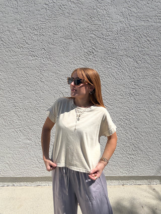 Woman wearing a white t-shirt and gray pants standing against a light gray wall.