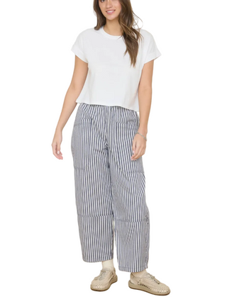 Woman wearing a white top and striped pants standing in a room with wooden flooring and a wall with decorative items.