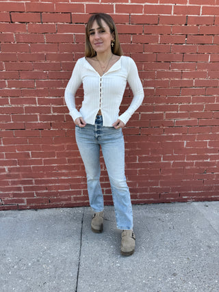 Woman wearing a white button-up shirt and light blue jeans standing against a red brick wall.