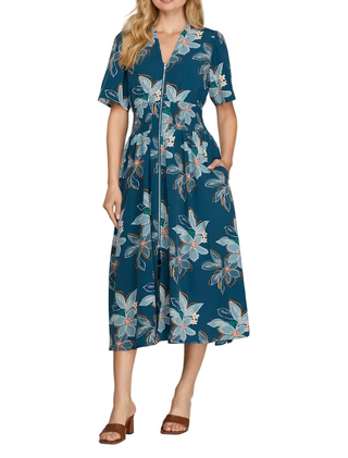 A woman models a short-sleeve midi dress with a teal blue background and a light blue, white, and orange floral print. The dress features a V-neckline with buttons, a fitted waist, and a flared skirt.
