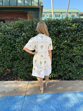 Woman in a floral dress walking past a hedge on a sunny day.
