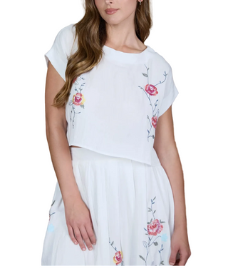 A woman wearing a white short-sleeve crop top and matching skirt with pink and yellow floral embroidery, posed in front of a brown striped wooden wall.