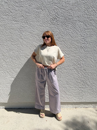 Woman wearing a white top and light purple pants against a gray wall.