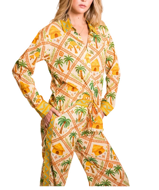 A woman modeling a matching long-sleeve button-up shirt and tie-waist pants set with a tropical print featuring palm trees, yellow houses, and orange geometric borders on a cream background.