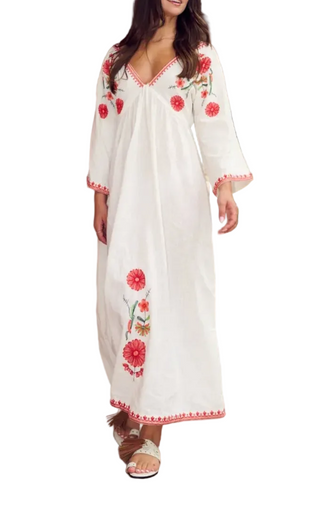A woman wearing a white maxi dress with red floral embroidery and red trim, walking in a covered outdoor corridor with potted plants.