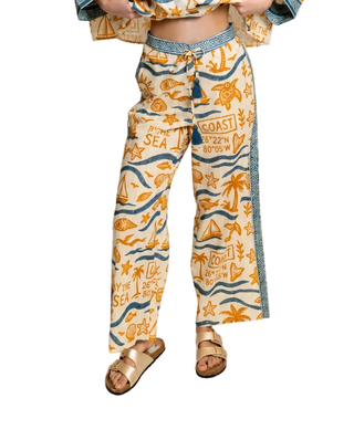 A person wearing wide-leg drawstring pants with a nautical print in orange and blue on a cream background, styled with metallic sandals against a plain white studio background.