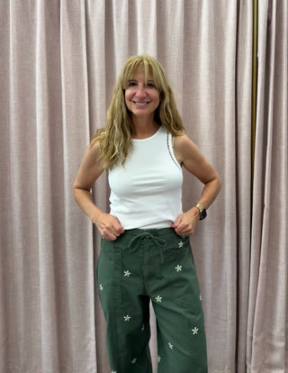 Woman wearing a white top and green pants with floral patterns against a beige curtain.