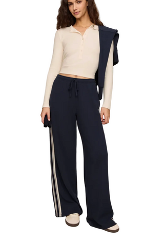 A model wearing a cream ribbed long-sleeve polo crop top and navy wide-leg pants with white side stripes, standing against a light beige studio background.