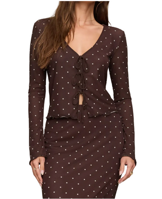 A woman wearing a dark brown long-sleeve dress with a white polka dot pattern, featuring a V-neckline with front tie closures and a matching skirt, set against a plain beige background.