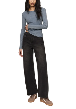 A model wearing a light blue long-sleeve knit top, black wide-leg jeans, and brown sneakers, standing against a plain beige studio background.