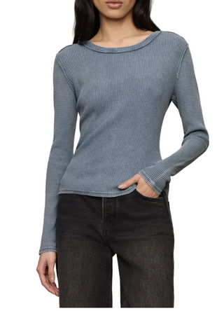 A woman wearing a fitted, long-sleeve ribbed knit top in a muted dusty blue color, paired with dark gray jeans, against a solid light beige background.