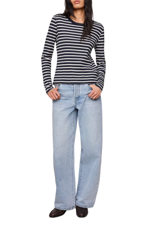 A model wearing a navy and white horizontally striped long-sleeve knit top with light-wash wide-leg jeans and dark brown shoes, standing against a solid beige studio background.