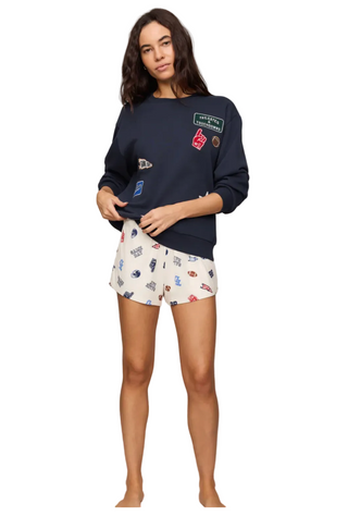 A woman modeling a navy blue crewneck sweatshirt with various colorful patches and white shorts featuring a colorful car pattern, set against a plain beige studio background.