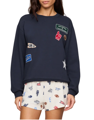 A woman wearing a navy blue crewneck sweatshirt with multiple colorful sports-themed embroidered patches, paired with white shorts featuring a football pattern, against a plain beige background. game day fit