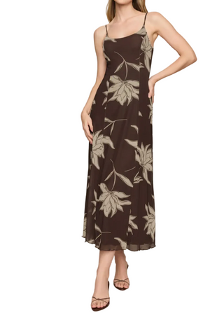 A woman modeling a brown spaghetti-strap midi dress with a beige leafy floral pattern, paired with brown strappy heels, against a solid light gray studio background.