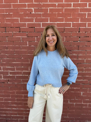 Woman wearing a light blue sweater and white pants standing against a red brick wall.