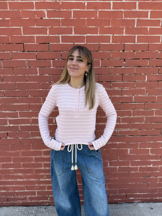 Woman wearing a pink striped sweater and blue jeans standing against a red brick wall.