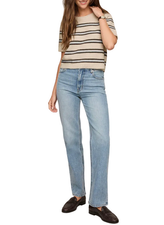 A woman modeling light-wash blue straight-leg jeans with a beige and black striped short-sleeve top and dark brown loafers, set against a plain beige studio background.