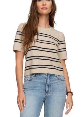 A woman wearing a beige short-sleeve knit sweater with thin black horizontal stripes and light blue jeans, shown from the shoulders down against a plain beige studio background.