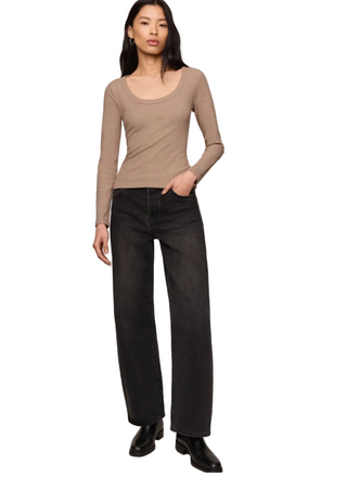 A model stands against a plain light beige studio background wearing a fitted long-sleeve scoop-neck knit top in taupe, dark charcoal wide-leg jeans, and black shoes.