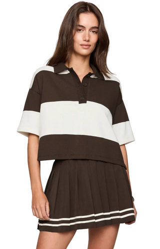 A woman modeling a short-sleeve color-block polo shirt and matching pleated mini skirt in dark brown and off-white, against a plain light beige studio background.
