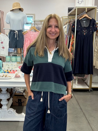 A woman stands in a boutique clothing store wearing a green and navy color-block short-sleeve polo shirt with white drawstrings and dark blue wide-leg denim pants.