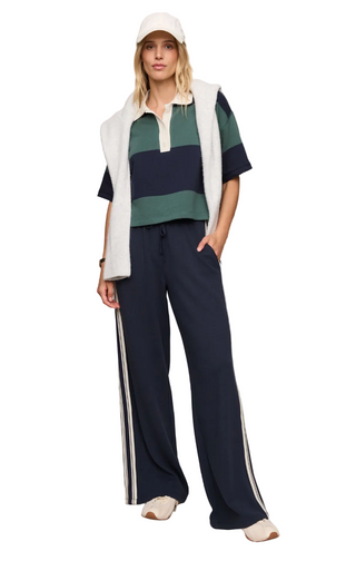 A woman modeling a coordinated outfit consisting of a navy and green striped short-sleeve polo shirt, a white sleeveless vest, navy wide-leg track pants with white side stripes, a white cap, and white slip-on shoes, against a plain beige studio background.