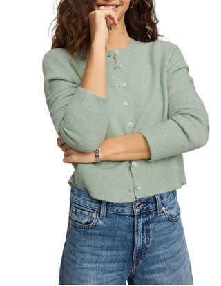 A woman wearing a light sage green knit cardigan with white buttons and blue jeans, posing against a neutral beige background.