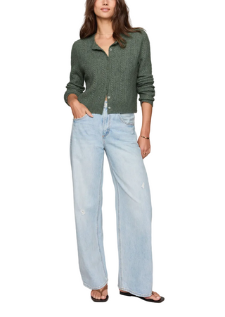 A woman modeling a dark green cable-knit cropped button-up cardigan with light-wash wide-leg denim jeans and black slide sandals, set against a plain beige studio background.