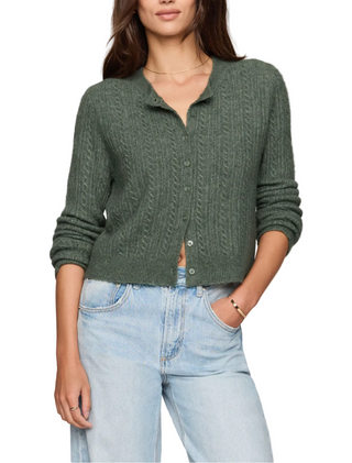 A woman wearing a sage green cable-knit button-up cardigan sweater and light-wash blue jeans, standing against a plain light beige studio background.