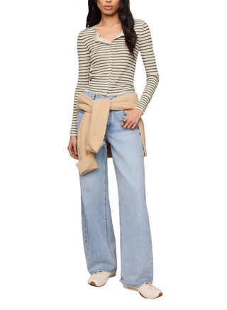 A model wearing a beige and black striped long-sleeve knit top, light-wash wide-leg jeans, and white slip-on shoes, with a beige cardigan tied around her waist, against a solid light beige studio background.