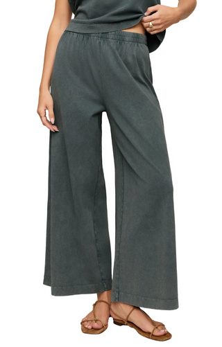 A person wearing green wide-leg pants with an elastic waistband, paired with brown strappy flat sandals, shown from the waist down against a plain beige studio background.