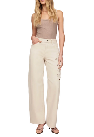 A model wearing off-white wide-leg pants with embroidered details on the side, paired with a taupe tank top and flat sandals, set against a plain light gray studio background.