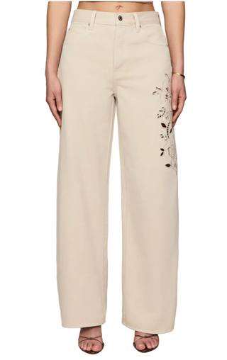 A model wearing a pair of wide-leg beige pants with black floral embroidery on the left leg, shown from the waist down on a white background.