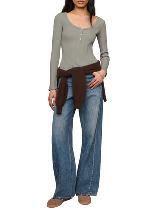 A model wearing a light sage green long-sleeve scoop-neck knit top with a button placket, styled with wide-leg blue jeans and a dark brown sweater tied around the waist, set against a plain beige studio background.