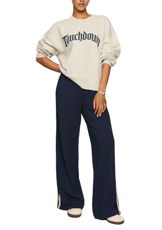 A model wearing a cream sweatshirt with 'Touchdown' printed in navy Gothic lettering, paired with navy wide-leg pants featuring white side stripes, set against a plain light gray studio background.