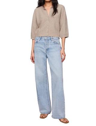 A model wearing a light beige cropped knit top and light-wash wide-leg blue jeans, standing against a solid beige studio background.