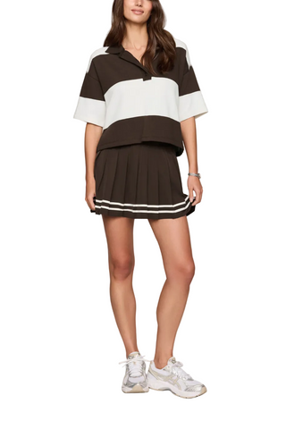 A woman models a short-sleeve collared knit top and matching pleated mini skirt set in dark brown and white, paired with white sneakers, against a plain light gray studio background.
