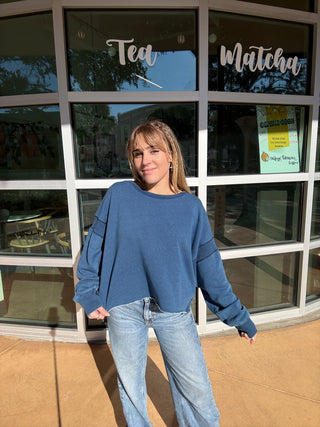 Person wearing a blue crewneck sweatshirt with cutout back and jeans standing in front of a store with large windows.
