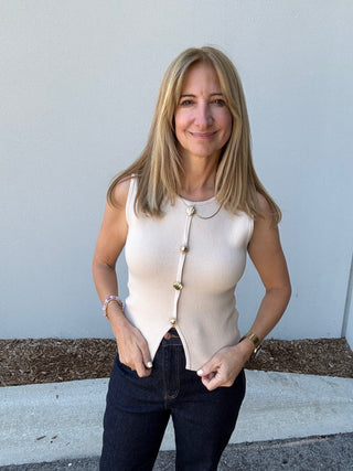 Woman wearing a sleeveless beige top and dark jeans standing against a light-colored wall.