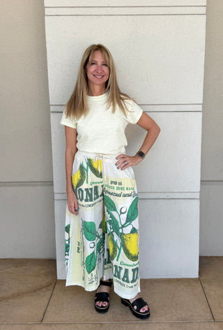Woman wearing a white top and colorful wide-leg pants with a lemon design, standing against a plain wall.