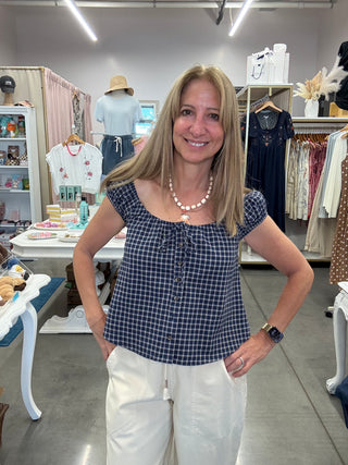 A woman with blonde hair stands in a clothing boutique wearing a navy and white gingham top with cap sleeves and a front tie detail, paired with white pants.