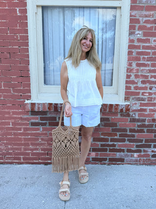 white pintuck top with no sleevesWoman standing in front of a brick wall holding a woven bag.
