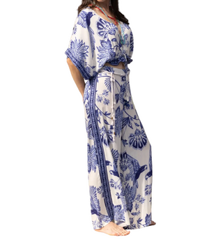 A woman wearing a white two-piece set with a blue tropical floral and bird print, standing barefoot on a wooden deck in front of a weathered gray wood wall.